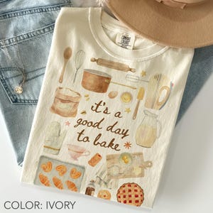 May include: Ivory-colored t-shirt with a watercolor illustration of baking tools and pastries. The text "it's a good day to bake" is featured on the front. The shirt is paired with blue jeans and a tan hat.