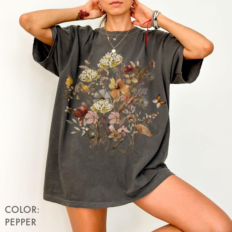 May include: Pepper-colored, oversized t-shirt featuring a floral design with a butterfly. The graphic includes a variety of flowers in shades of yellow, pink, and brown. The shirt has short sleeves and a relaxed fit.