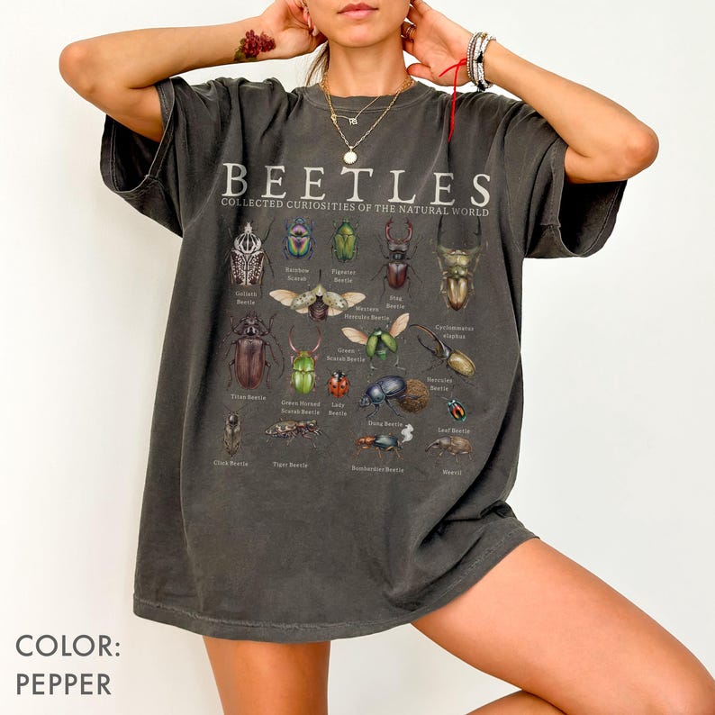 Puede incluir: Camiseta color pimienta con la palabra "BEETLES" e ilustraciones de insectos. El texto "COLLECTED CURIOSITIES OF THE NATURAL WORLD" est&aacute; sobre las im&aacute;genes de insectos. La camiseta es de corte holgado.