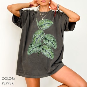以下が含まれることがあります： ペッパーカラーの半袖Tシャツで、中央に緑と銀の斑が入ったハート型の葉を持つつる性植物のグラフィックイラストが描かれています。ゆったりとしたフィット感のTシャツで、クルーネックの襟元が特徴的なボタニカルデザインです。