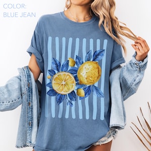 May include: Blue jean colored t-shirt with a lemon graphic. The graphic features a sliced lemon, a whole lemon, and blue leaves. Vertical light blue stripes are behind the lemons. The text "COLOR: BLUE JEAN" is at the top left.