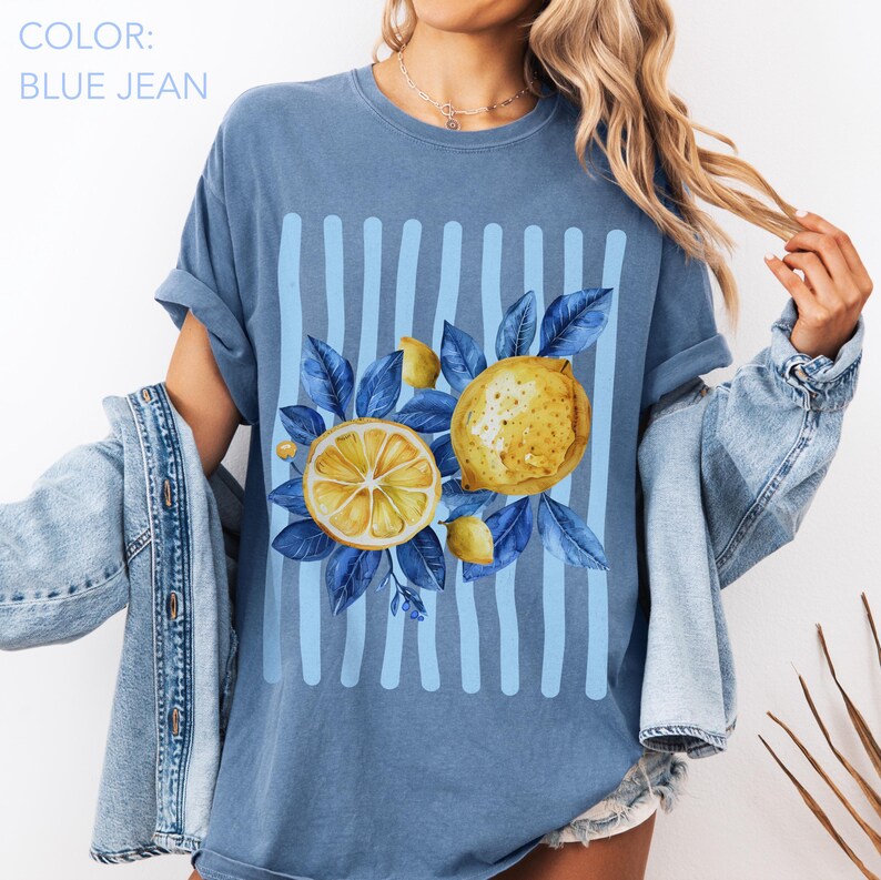 May include: Blue jean colored t-shirt with a lemon graphic. The graphic features a sliced lemon, a whole lemon, and blue leaves. Vertical light blue stripes are behind the lemons. The text "COLOR: BLUE JEAN" is at the top left.