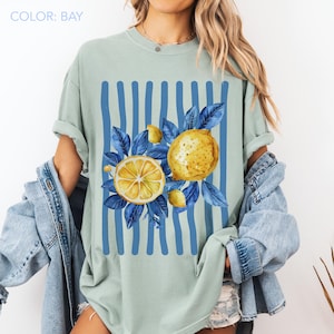 May include: A light green t-shirt with a lemon graphic. The design features a sliced lemon, whole lemons, and blue leaves against a backdrop of blue vertical stripes. The text "COLOR: BAY" is at the top.