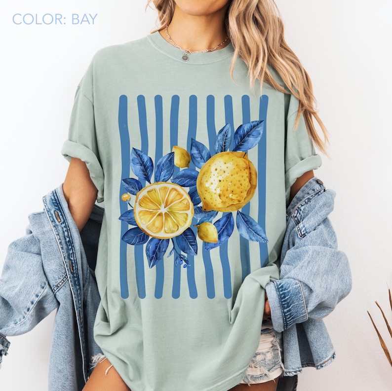 May include: A light green t-shirt with a lemon graphic. The design features a sliced lemon, whole lemons, and blue leaves against a backdrop of blue vertical stripes. The text "COLOR: BAY" is at the top.