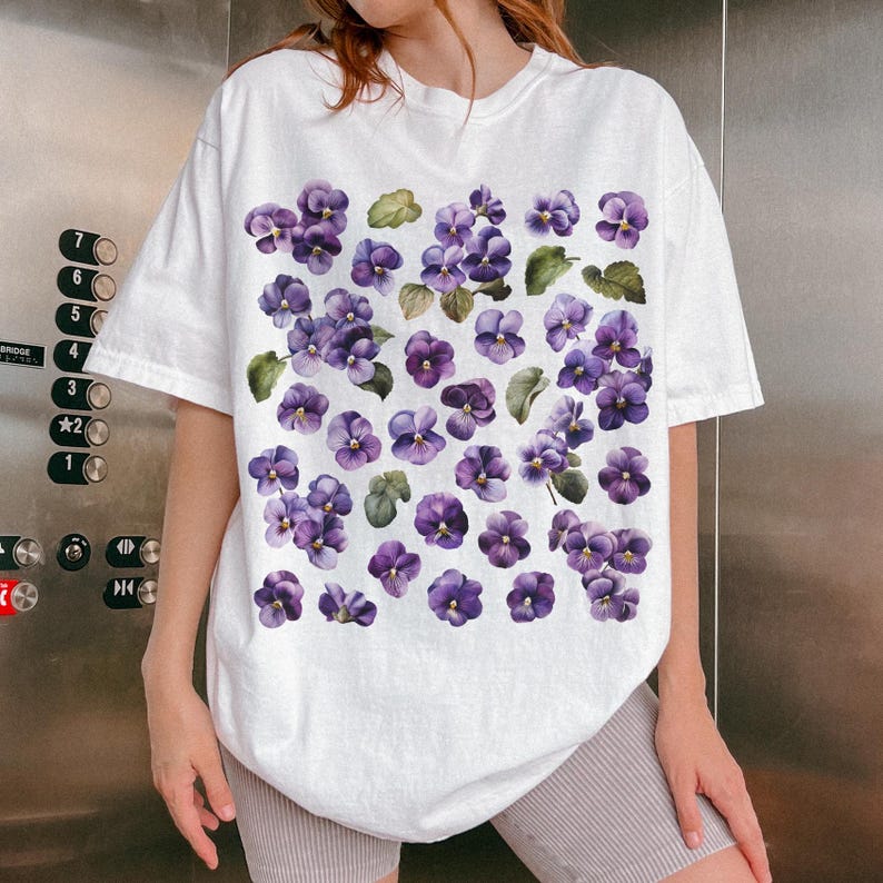 May include: White t-shirt featuring a pattern of purple violets and green leaves. The floral design is printed across the front of the shirt. The shirt has short sleeves and a relaxed fit.