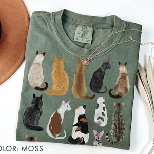 May include: Moss green t-shirt featuring a colorful illustration of various cat breeds. The shirt has a crew neck and short sleeves. A silver necklace with a pendant is draped across the shirt. The text "COLOR: MOSS" is at the bottom.