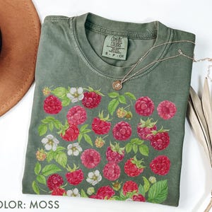 Op de afbeelding: Een mosgroen t-shirt met een levendige frambozen- en bloemenprint. Het ontwerp toont gedetailleerde illustraties van rode frambozen, witte bloemen en groene bladeren. Een delicate gouden ketting rust op het shirt. Het label luidt "Comfort Colors".