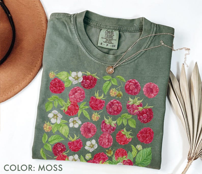 Op de afbeelding: Een mosgroen t-shirt met een levendige frambozen- en bloemenprint. Het ontwerp toont gedetailleerde illustraties van rode frambozen, witte bloemen en groene bladeren. Een delicate gouden ketting rust op het shirt. Het label luidt "Comfort Colors".
