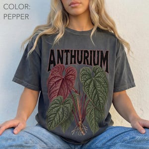 May include: A pepper-coloured short-sleeve t-shirt featuring a graphic of an Anthurium plant with large red and green heart-shaped leaves. The word ANTHURIUM is printed in a bold, dark, arched font above the botanical illustration of the plant.
