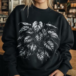 Peut inclure: Sweat-shirt noir orné d'une illustration botanique blanche d'une plante aux grandes feuilles à motifs. Le motif est centré sur le devant. L'arrière-plan est flou, mettant en valeur le graphisme. Le sweat-shirt est de style décontracté et confortable.