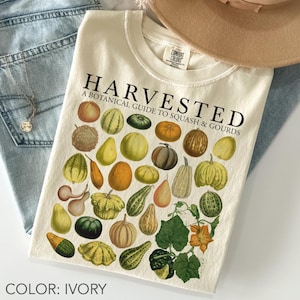 Puede incluir: Camiseta de color marfil con la palabra "HARVESTED" y una guía botánica de calabazas. La camiseta muestra ilustraciones de varios tipos de calabazas en tonos verdes, naranjas, amarillos y marrones. La camiseta está doblada sobre unos vaqueros azules.