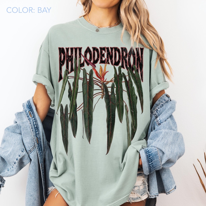 May include: A bay-colored t-shirt featuring a graphic print of dark green philodendron leaves and a central orange flower. The word PHILODENDRON is printed in a bold, distressed black font above the botanical illustration on the front of the shirt.