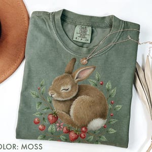 Puede incluir: Una camiseta verde musgo con una ilustración detallada de un conejo marrón rodeado de fresas y hojas. La camiseta tiene cuello redondo y mangas cortas. Un collar plateado con un pequeño colgante descansa sobre la camiseta.