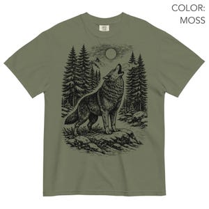 Puede incluir: Una camiseta de manga corta color verde musgo con un gr&aacute;fico en tinta negra de un lobo aullando a la luna llena. La escena incluye un bosque de pinos, terreno rocoso y monta&ntilde;as al fondo. El texto COLOR: MOSS es visible en la imagen.