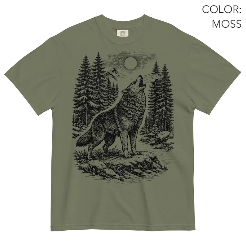 Puede incluir: Una camiseta de manga corta color verde musgo con un gr&aacute;fico en tinta negra de un lobo aullando a la luna llena. La escena incluye un bosque de pinos, terreno rocoso y monta&ntilde;as al fondo. El texto COLOR: MOSS es visible en la imagen.