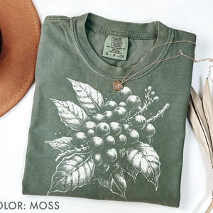Puede incluir: Una camiseta de color musgo con una ilustración blanca de bayas y hojas de café. Un collar dorado con un colgante descansa sobre la camiseta. La camiseta es de la marca Comfort Colors, talla S-CH.