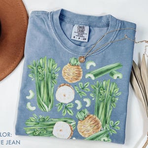 May include: Blue jean colored t-shirt with a watercolor design of celery and celeriac. The shirt has a crew neck and short sleeves. A gold necklace with a pendant is draped across the shirt.