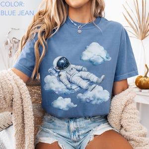 Peut inclure: T-shirt bleu jean avec une illustration &agrave; l'aquarelle d'un astronaute se relaxant sur un nuage. L'astronaute porte une combinaison spatiale et un casque blancs. Le t-shirt a un col rond et des manches courtes. Le texte "COLOR: BLUE JEAN" est en haut &agrave; gauche.