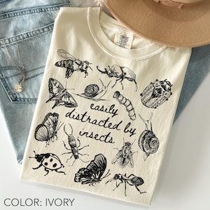 Op de afbeelding: Een ivoorkleurig t-shirt met zwarte lijntekeningen van diverse insecten, waaronder vlinders, kevers, slakken en een bij. De tekst "easily distracted by insects" is in het midden gedrukt in een cursief lettertype.