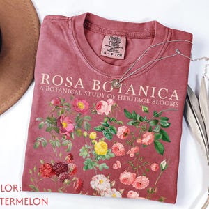 Könnte beinhalten: Ein wassermelonfarbenes T-Shirt mit dem Text ROSA BOTANICA A BOTANICAL STUDY OF HERITAGE BLOOMS in cremefarbener Schrift. Das T-Shirt zeigt eine detaillierte Grafik verschiedener Rosen in Rosa-, Gelb-, Rot- und Weißtönen mit grünen Blättern.