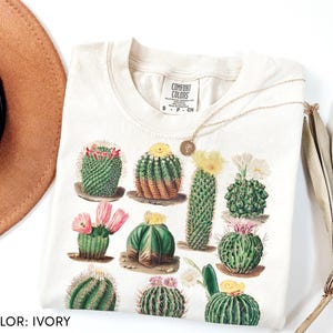 Op de afbeelding: Een ivoorkleurig t-shirt met een grafische print van diverse groene cactussen met roze en gele bloemen. Het t-shirt heeft een Comfort Colors-merklabel bij de binnenhalslijn, wat de maat small aangeeft. Het ontwerp toont meerdere cactussoorten.