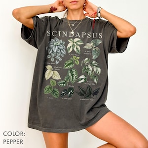 以下が含まれることがあります： ペッパーカラーのオーバーサイズTシャツ。様々なスキンダプサス植物の品種を描いたボタニカルグラフィックプリントが施されています。デザインには葉の詳細なイラストが含まれており、各植物のイラストの下には白い文字で学名が印刷されています。