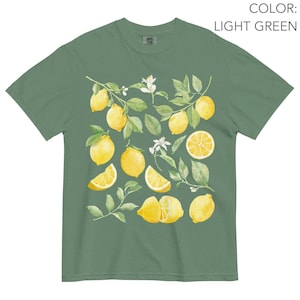 May include: Light green t-shirt featuring a watercolor design of lemons, leaves, and blossoms. The lemons are yellow, with some sliced to show the inside. The leaves are green, and the blossoms are white. The shirt is a comfortable, casual style.