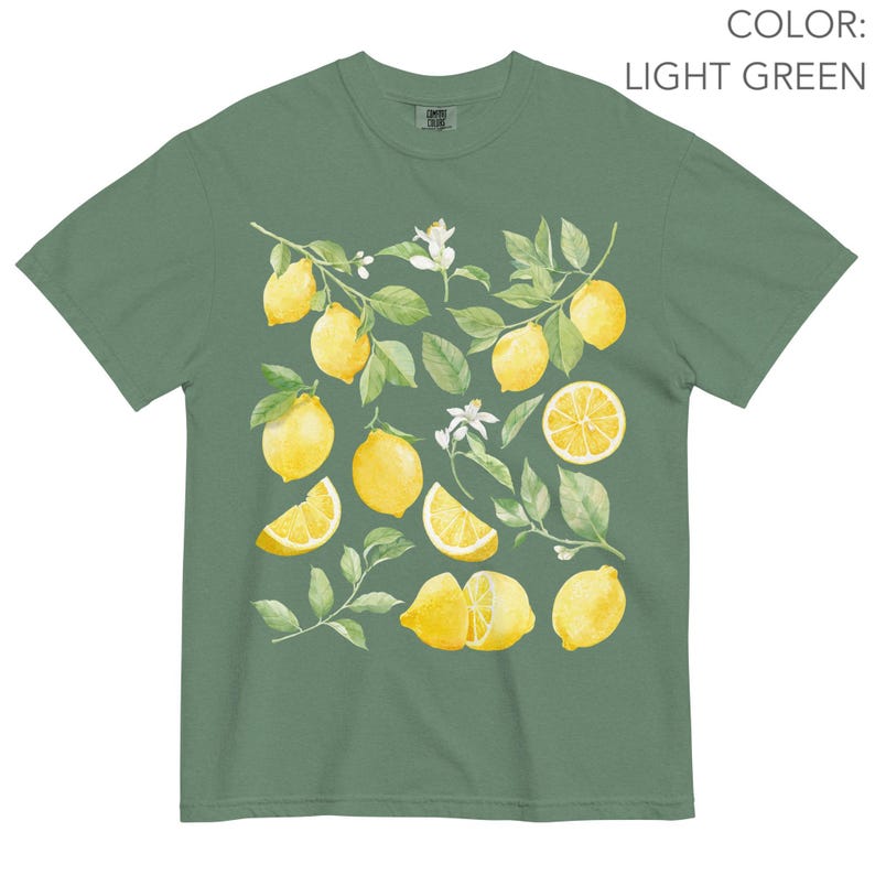 May include: Light green t-shirt featuring a watercolor design of lemons, leaves, and blossoms. The lemons are yellow, with some sliced to show the inside. The leaves are green, and the blossoms are white. The shirt is a comfortable, casual style.