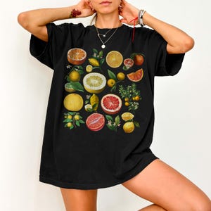 May include: A black short-sleeve t-shirt featuring a vibrant graphic print of various citrus fruits including oranges, grapefruits, lemons, and limes, displayed in whole and sliced forms with green leaves and small white blossoms scattered throughout.