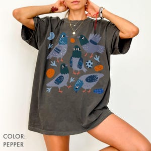 May include: Pepper-colored t-shirt featuring a whimsical illustration of several pigeons in various poses. The design includes blue, gray, and orange accents, with additional graphic elements. The shirt has a relaxed fit.