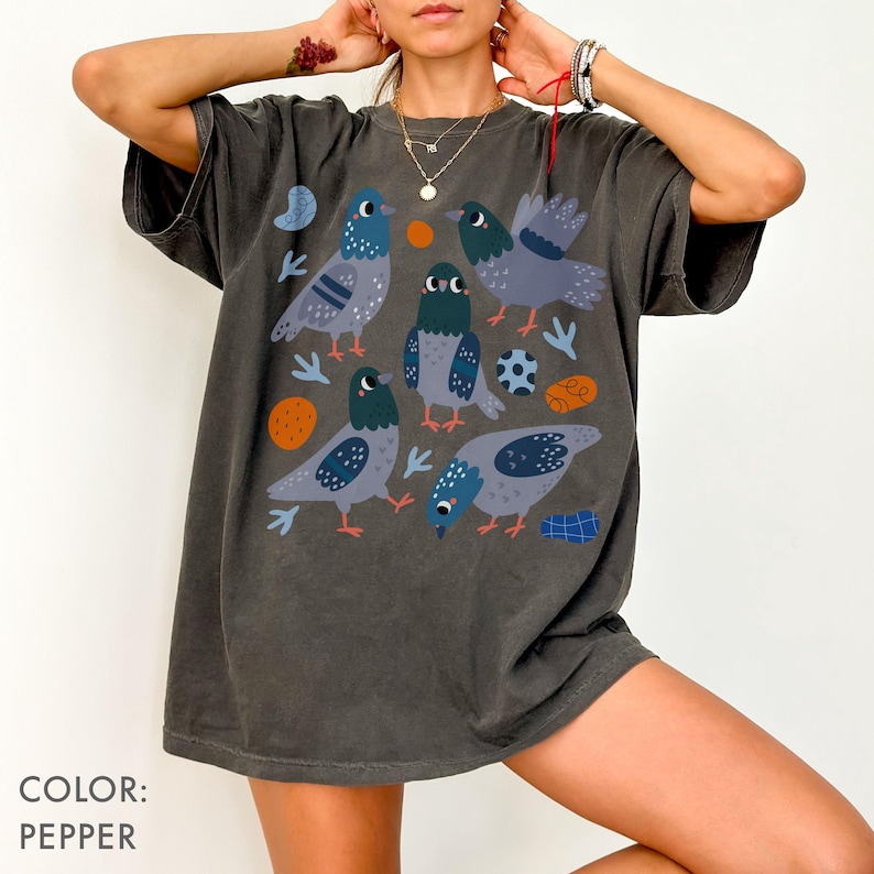 May include: Pepper-colored t-shirt featuring a whimsical illustration of several pigeons in various poses. The design includes blue, gray, and orange accents, with additional graphic elements. The shirt has a relaxed fit.
