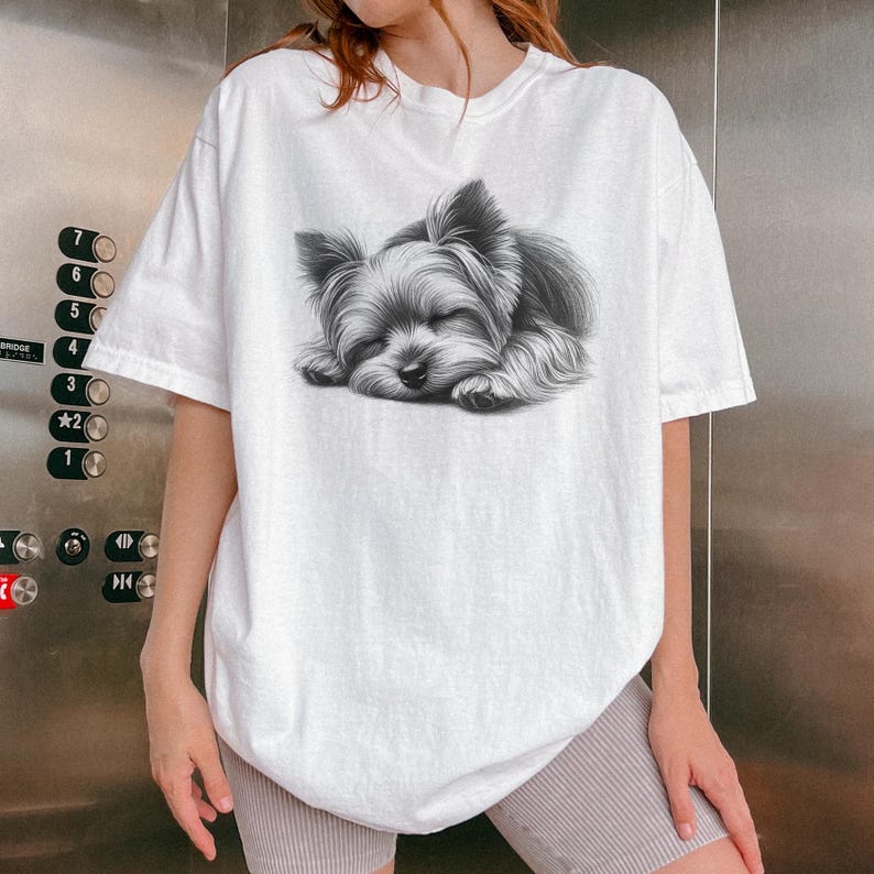 Puede incluir: Camiseta blanca con una ilustraci&oacute;n en blanco y negro de un Yorkshire Terrier durmiendo. El perro est&aacute; representado con los ojos cerrados, descansando sobre sus patas. La camiseta tiene un corte holgado y mangas cortas.