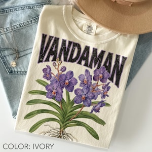 May include: Ivory-colored t-shirt with the word "VANDAMAN" in black, above a purple orchid illustration. The shirt is folded on top of blue jeans, with a gold necklace and a tan hat visible.