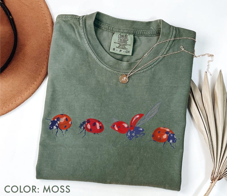 May include: Moss green t-shirt with a ladybug design. The shirt has a crew neck and short sleeves. A gold necklace with a small pendant is draped across the shirt. The shirt is made of a soft, comfortable material.