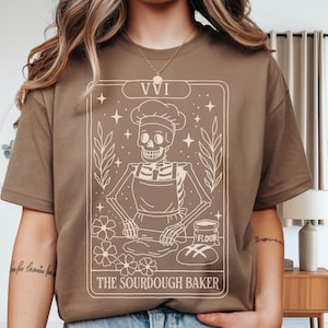 May include: A brown t-shirt featuring a tarot card design with a skeleton baker wearing a chef's hat and apron. The card reads "VVI" at the top and "The Sourdough Baker" at the bottom. The design includes stars, flowers, and leaves.