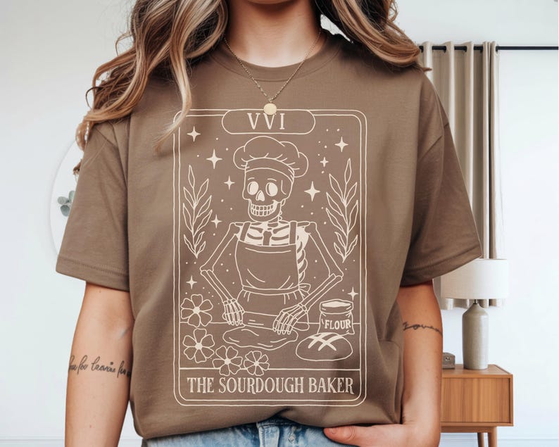 May include: A brown t-shirt featuring a tarot card design with a skeleton baker wearing a chef's hat and apron. The card reads "VVI" at the top and "The Sourdough Baker" at the bottom. The design includes stars, flowers, and leaves.