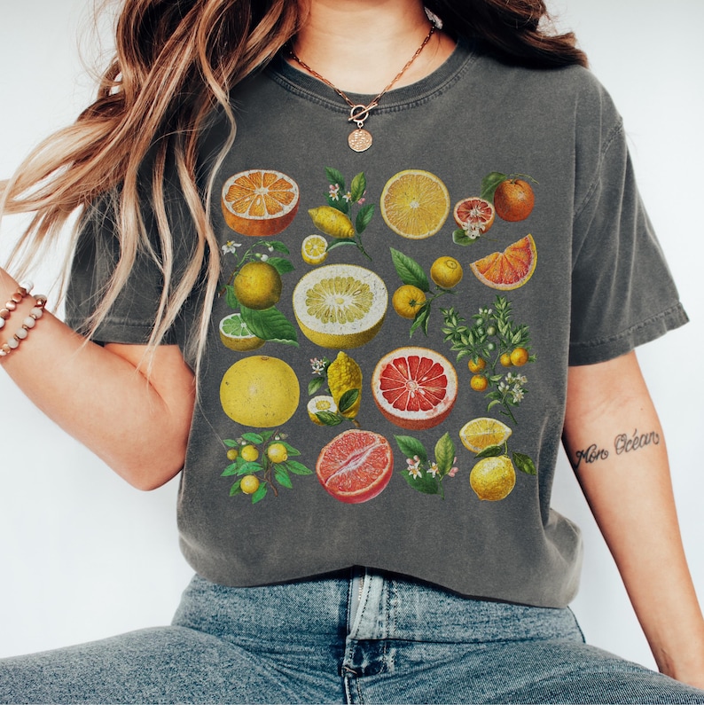 May include: A charcoal gray short-sleeve t-shirt featuring a botanical print of various citrus fruits including oranges, grapefruits, lemons, and limes, displayed as whole fruits, sliced halves, and small leafy branches with blossoms.