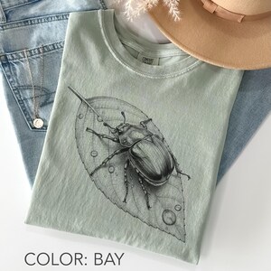 Op de afbeelding: Lichtgroen t-shirt met een gedetailleerde zwart-wit illustratie van een kever op een blad met waterdruppels. De tekst "COLOR: BAY" staat onderaan. Het shirt ligt op een blauwe jeans.