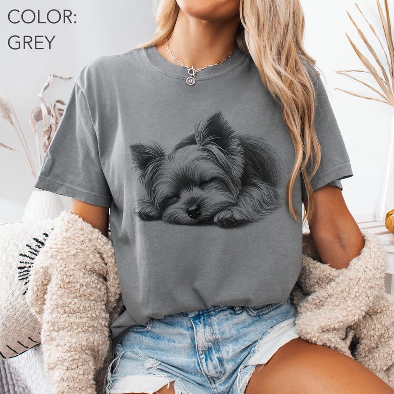 Puede incluir: Camiseta gris con una ilustraci&oacute;n en blanco y negro de un Yorkshire Terrier durmiendo. El gr&aacute;fico detallado contrasta con la tela gris. La camiseta tiene cuello redondo y mangas cortas, un estilo casual.