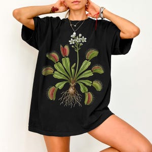 May include: Black t-shirt featuring a botanical illustration of a Venus flytrap. The graphic includes green leaves, red-lined traps, white flowers, and detailed roots. The shirt is a comfortable, oversized fit.