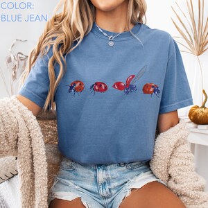 May include: Blue jean coloured t-shirt featuring a ladybug design. The graphic shows four ladybirds in different poses, with red and black details. The shirt has short sleeves and a crew neck.