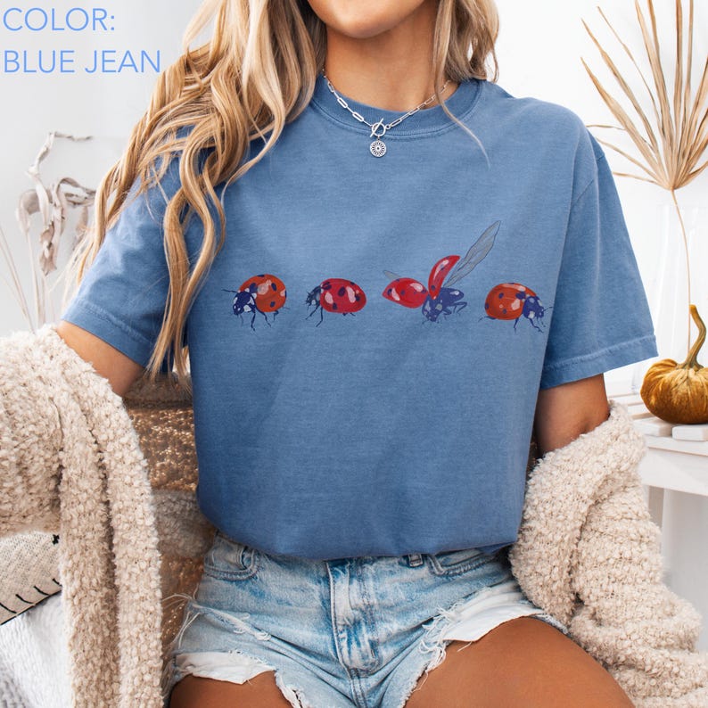 May include: Blue jean coloured t-shirt featuring a ladybug design. The graphic shows four ladybirds in different poses, with red and black details. The shirt has short sleeves and a crew neck.
