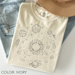 May include: Ivory-colored t-shirt featuring a black line drawing of a solar system with planets, stars, and a sun. The shirt is folded on top of blue jeans, with a gold necklace and a tan hat in the background. The text "COLOR: IVORY" is at the bottom.