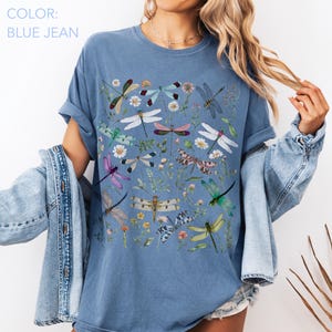 May include: Blue jean colored t-shirt featuring a colorful dragonfly and floral design. The shirt has a crew neck and short sleeves. The text "COLOR: BLUE JEAN" is in the upper left corner.