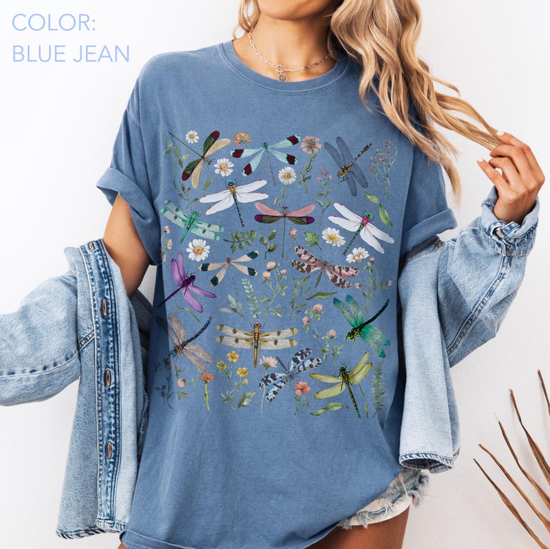 May include: Blue jean colored t-shirt featuring a colorful dragonfly and floral design. The shirt has a crew neck and short sleeves. The text "COLOR: BLUE JEAN" is in the upper left corner.