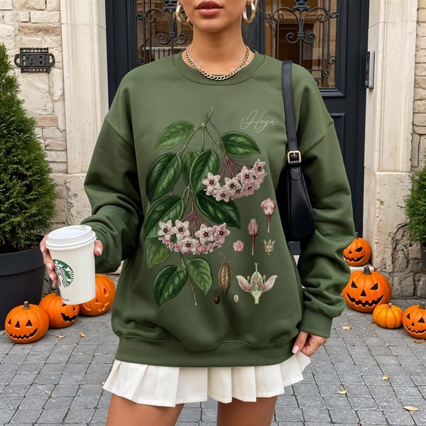 May include: Olive green sweatshirt featuring a botanical illustration of a Hoya plant with pink flowers and green leaves. The word "Hoya" is written in script. The sweatshirt is paired with a white pleated skirt.