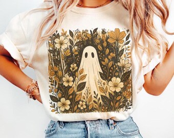 Op de afbeelding: Crèmekleurig T-shirt met een spookontwerp. Het spook is omgeven door een bloemstuk met beige en bruine bloemen en bladeren. Het shirt wordt gecombineerd met een blauwe spijkerbroek.
