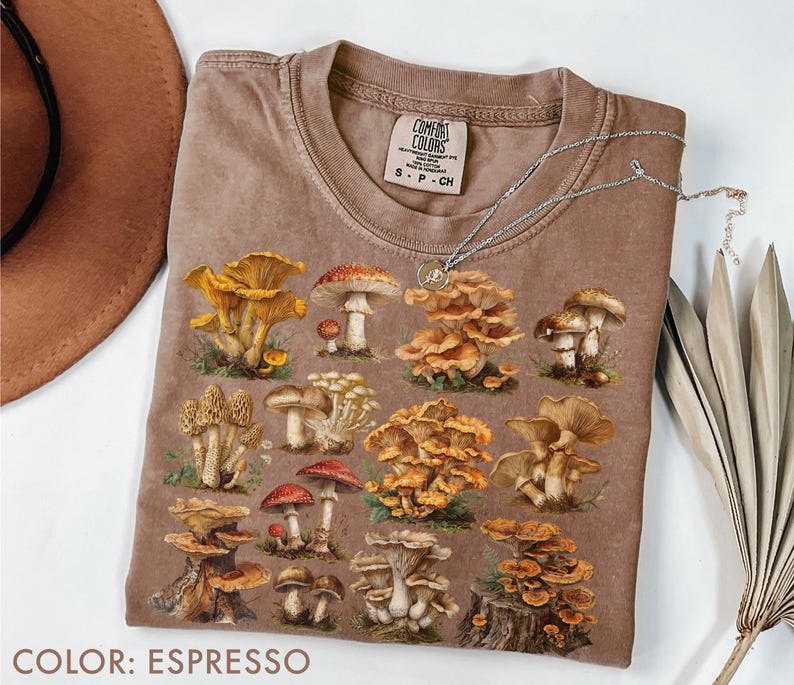 Pu&ograve; includere: T-shirt color espresso con illustrazioni di vari funghi. La maglietta ha un girocollo e maniche corte. Una collana d'argento con un piccolo ciondolo &egrave; appoggiata sulla maglietta. Il testo "COLOR: ESPRESSO" &egrave; in basso nell'immagine.