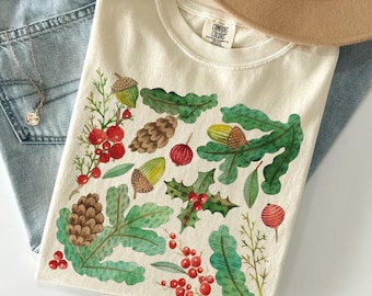 May include: Ivory-colored t-shirt featuring a watercolor design of acorns, pinecones, holly, and berries. The shirt is folded on top of blue jeans, with a necklace and a tan hat visible in the background. The text "COLOR: IVORY" is at the bottom.