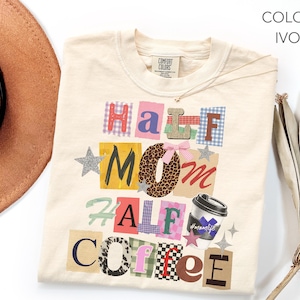 May include: Ivory-colored t-shirt with the words "Half Mom Half Coffee" in a collage style. The design includes a coffee cup graphic and star accents. The shirt is made of a soft material and is a Comfort Colors brand.